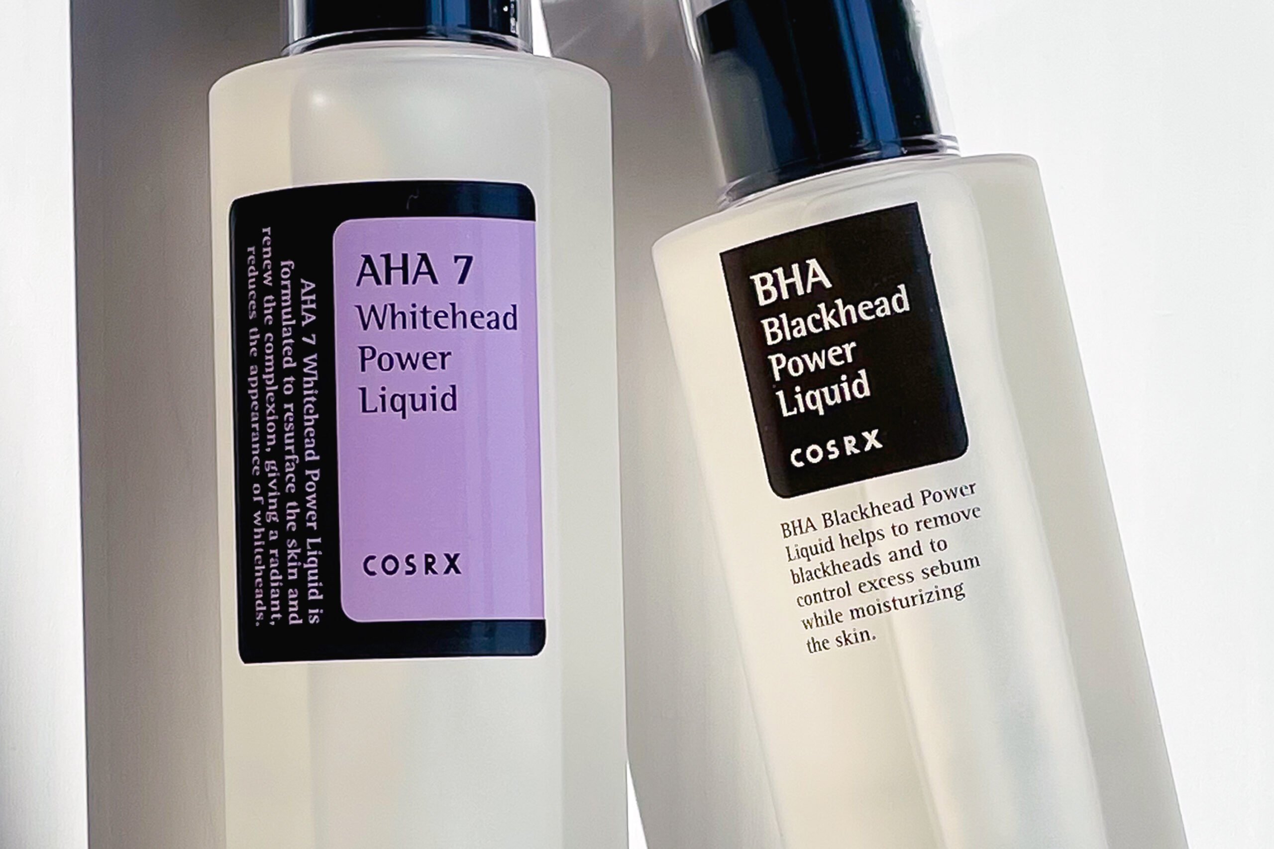 The image showcases two skincare products from the brand COSRX, positioned side-by-side on a reflective white surface. Both products are packaged in minimalist, clear plastic bottles with black caps and labels. The bottle on the left features a bright green label with white text, identifying it as “AHA 7 Whitehead Power Liquid.” The label details that this liquid is formulated to resurface the skin, renew complexion, and reduce the appearance of whiteheads, aiming for a radiant finish. The bottle on the right has a black label with white text, labeled “BHA Blackhead Power Liquid.” Its description states that it helps remove blackheads and control excess sebum while simultaneously moisturizing the skin. Both labels prominently display the COSRX logo within a rectangular white box. The bottles are slightly angled, allowing a clear view of the liquid inside – the AHA liquid appears clear, while the BHA liquid seems slightly more viscous. The reflective surface creates subtle highlights and shadows, emphasizing the bottle shapes and label details. The overall aesthetic is clean, modern, and emphasizes the functional benefits of the skincare products. The lighting is soft and diffused, contributing to a professional product shot feel. The composition suggests a comparison between the two products, highlighting their different active ingredients (AHA and BHA) and targeted concerns (whiteheads vs. blackheads).